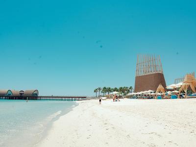 Weißer Sandstrand mit klarem Wasser, Palmen und modernen Hotelgebäuden unter blauem Himmel.