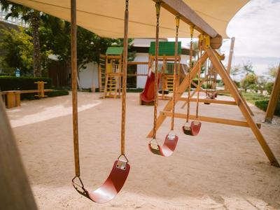Spielplatz mit Schaukeln und Sand unter einem Sonnensegel, umgeben von Bäumen und Gartenmöbeln.