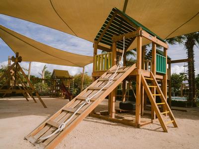 Kinderspielplatz mit Holzklettergerüst und Sonnensegeln neben Palmen im Hotelgarten.