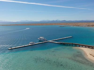 Langer Steg erstreckt sich über klares türkisfarbenes Wasser zu einem weißen Sandstrand unter blauem Himmel.