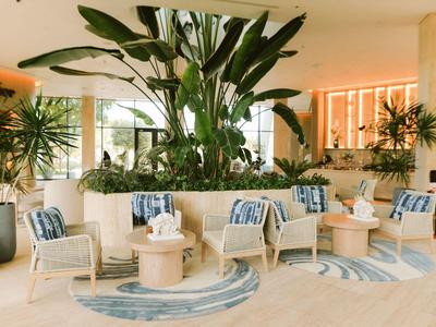 Hell eingerichteter Hotel-Loungebereich mit Pflanzen, bequemen Sesseln und runden Tischen.