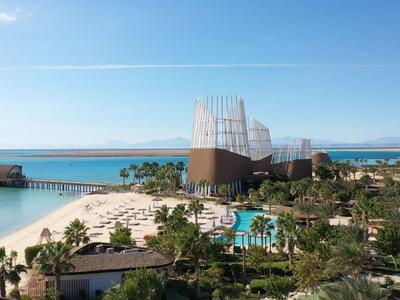 Moderner Hotelkomplex am Meer mit Pool, Palmen und Strand unter klarem blauem Himmel.