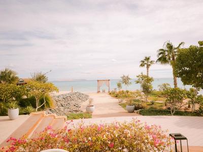 Blick auf einen Weg mit Blumen und Palmen, der zu einer Strandansicht mit einem Pavillon führt.