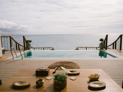 Blick auf einen Infinity-Pool mit Meerblick von einer Terrasse mit Tisch und Pflanzen.