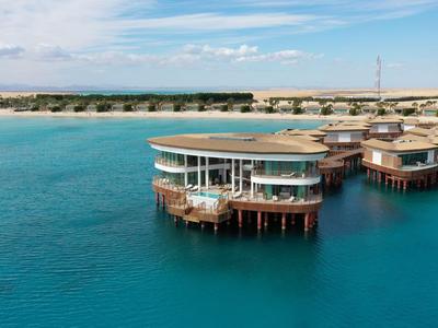 Runde Bungalows auf Stelzen über klarem, türkisfarbenem Wasser mit Sandstrand im Hintergrund.