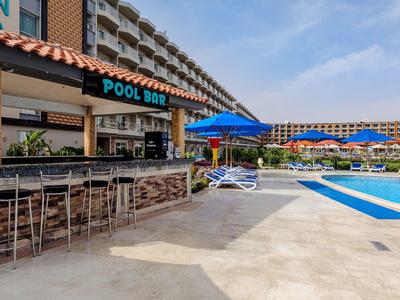 Poolbar mit Hockern neben großem Schwimmbad und Sonnenschirmen vor einem Hotelgebäude.