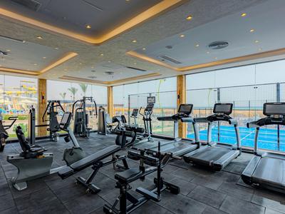 Helles Fitnessstudio mit verschiedenen Trainingsgeräten und Ausblick auf den Poolbereich.