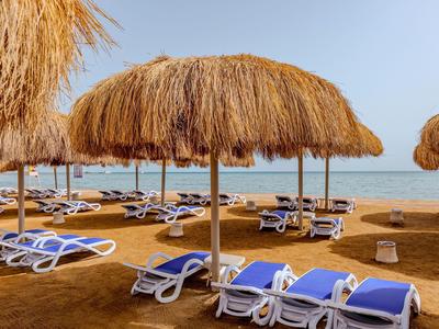 Strand mit blauen Liegestühlen und Strohmatschirmen unter klarem Himmel.