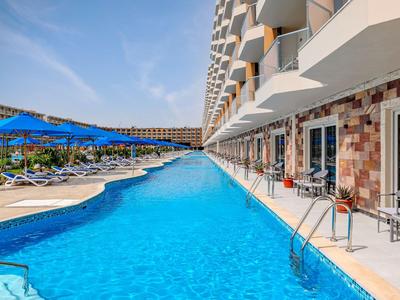 Moderner Hotelpool mit blauem Wasser und Liegestühlen unter Sonnenschirmen an einem sonnigen Tag.
