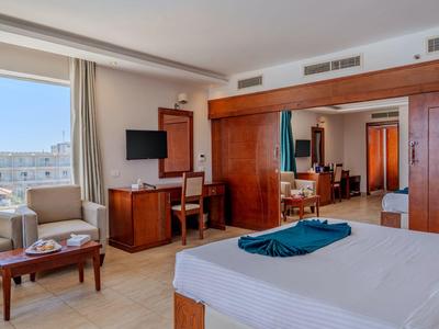 Hell eingerichtetes Hotelzimmer mit großem Bett, Sofa, Fernseher und Blick auf Stadt.