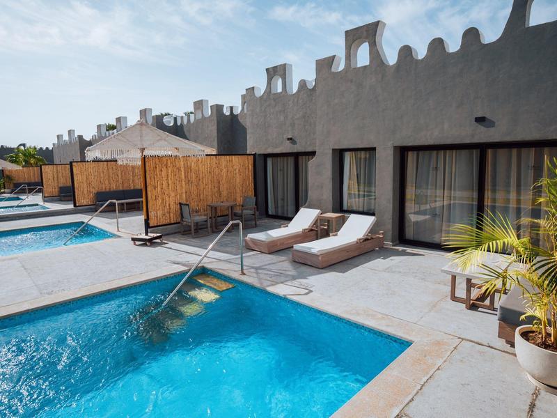 Poolside lounge area with sunbeds and doors leading to rooms under a cloudy blue sky.