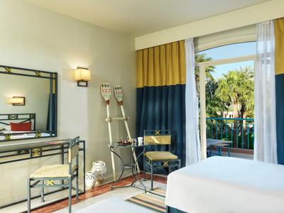 Hotelový pokoj s manželskou postelí, zrcadlem, židlemi a balkonem s výhledem na palmy.