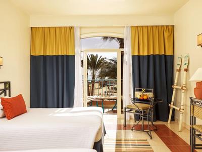 Světlý hotelový pokoj s manželskou postelí, závěsy a výhledem na terasu s palmami.