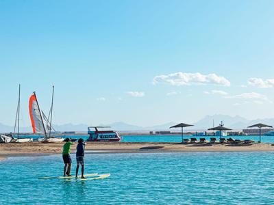 Dva lidé stojí na paddleboardu v klidném moři u pláže s deštníky a loděmi.