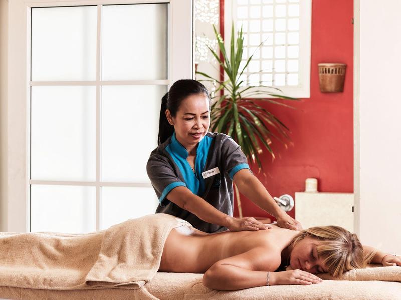 Hotelová pracovnice masíruje uvolněného hosta v světlé místnosti s rostlinami.