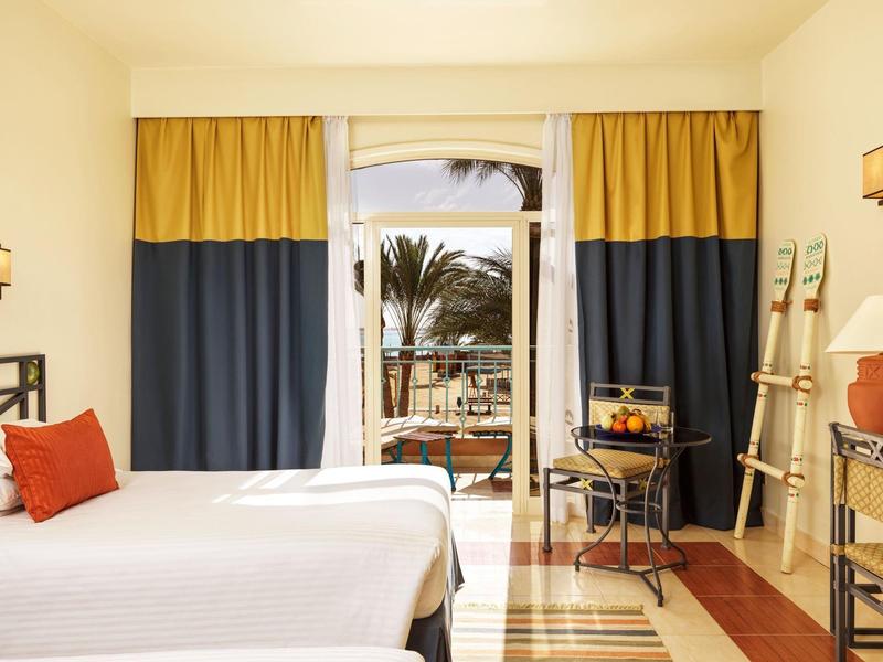 Světlý hotelový pokoj s manželskou postelí, závěsy a výhledem na terasu s palmami.