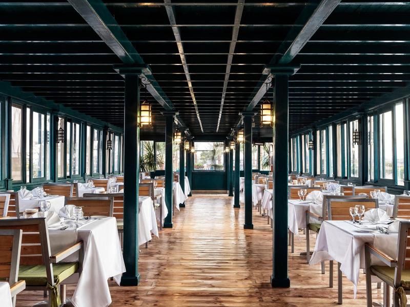 Velká jídelna s dřevěnou podlahou, bílými ubrusy a mnoha okny z obou stran.