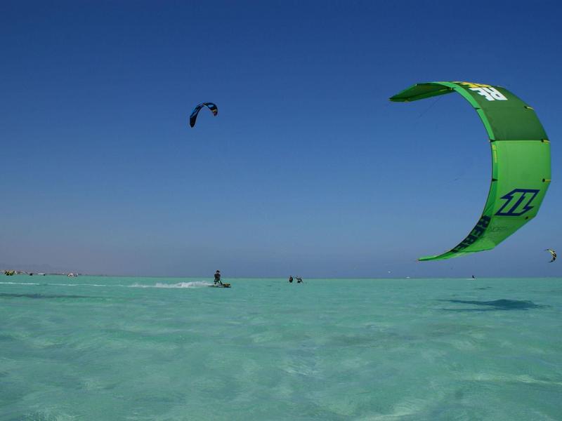 Zelený kitesurfový drak nad čistou, mělčí vodou pod modrou oblohou na pláži.