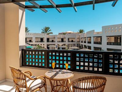 Balkon met rieten stoelen en tafel met tropisch drankje, uitzicht op hotel en zwembad.