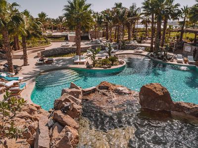 Luxury hotel pool surrounded by palm trees, sun loungers, and rock formations under clear weather.