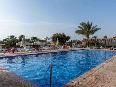 Large outdoor pool with sun loungers and umbrellas in a hotel setting under a blue sky.