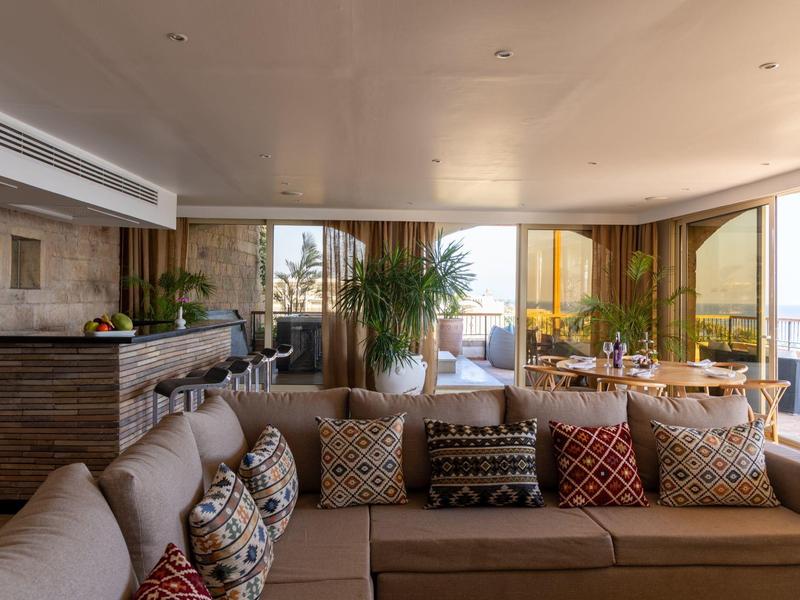 Przytulny salon z dużymi sofami i wieloma poduszkami, panoramiczne okna na balkon z roślinami.