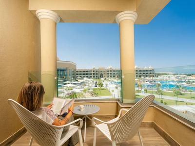 Balcony with two chairs and table overlooking hotel pool and buildings under a blue sky.