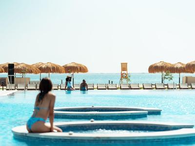Woman sitting at pool edge with sun loungers and umbrellas by the sea in background.