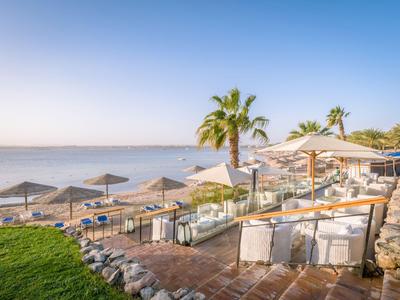 Seating area on terrace overlooking sandy beach, sun umbrellas, and calm sea under clear blue sky.