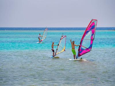 Tři lidé windsurfují na čisté modré vodě pod zataženou oblohou.