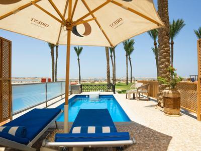Zwembad met blauwe ligstoel en parasol naast palmbomen en zeezicht bij hotel.