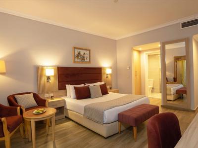 Lichte hotelkamer met tweepersoonsbed, twee fauteuils, houten meubels en zachte verlichting.