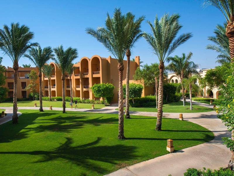 Teren hotelu z zadbanym trawnikiem, palmami i beżowymi budynkami pod niebieskim niebem.