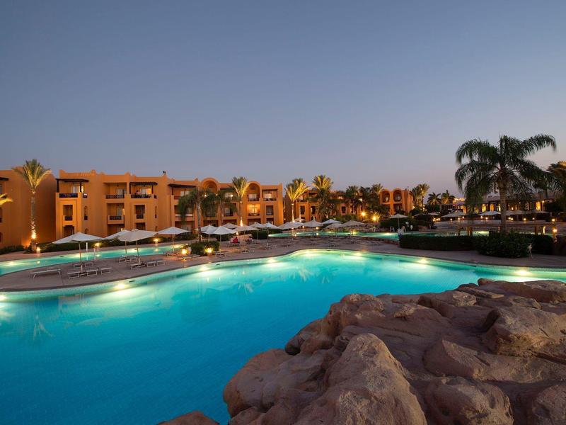 Oświetlony basen z palmami i budynkami hotelu o zmierzchu.