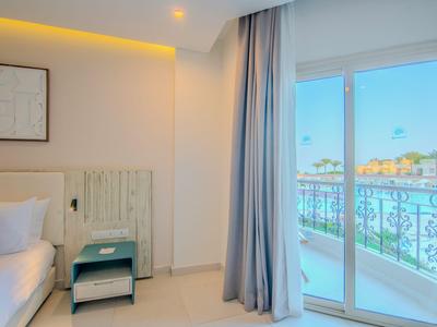 Helles Hotelzimmer mit Bett, Nachttisch und Blick auf einen Balkon mit Geländer.