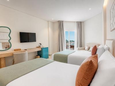 Bright hotel room with two double beds, TV, desk, and window with outside view.
