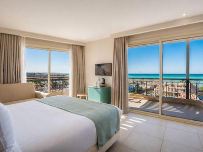 Bright hotel room with double bed, balcony access, sea view, and cityscape.