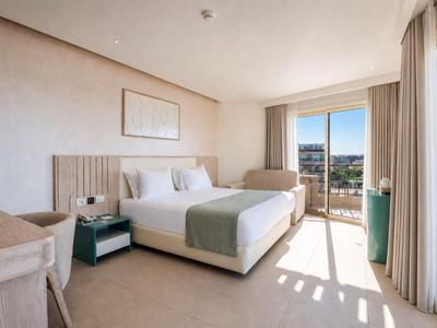 Bright hotel room with double bed, armchair, desk, and balcony overlooking the sea.