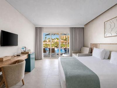 Bright hotel room with double bed, desk, TV, and view of pool and houses.