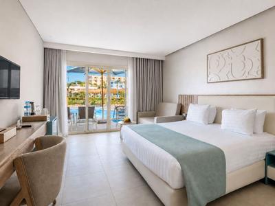 Bright hotel room with large bed, desk, balcony, and view of a hotel complex.