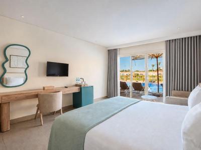 Bright hotel room with large bed, desk, TV, and balcony overlooking palm trees.
