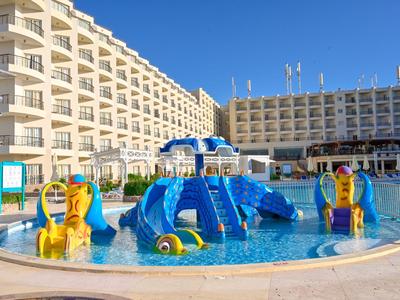 Un hotel con piscina al aire libre y área de juegos infantil colorida en el agua bajo el cielo azul.