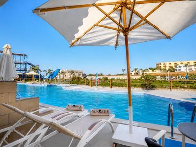 Pool area with lounge chairs and umbrellas in front of a hotel complex under a blue sky.