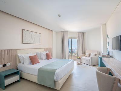 Habitación de hotel luminosa con cama doble, zona de estar, escritorio y balcón con vista al mar.