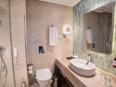 Baño moderno de hotel con lavabo, inodoro, ducha y gran espejo iluminado.