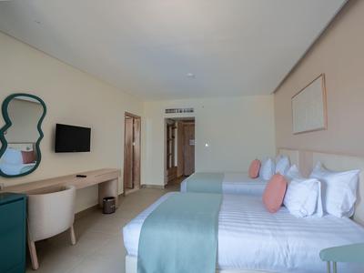 Habitación de hotel luminosa con dos camas, mantas azul-verde, cojines rosas y área de trabajo moderna.