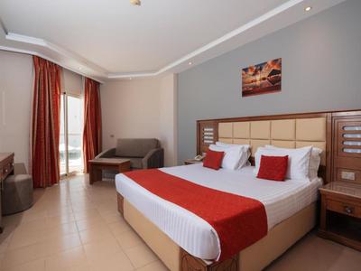 Modernes Hotelzimmer mit Doppelbett, roter Bettdecke, Vorhängen und Sitzbereich neben dem Fenster.