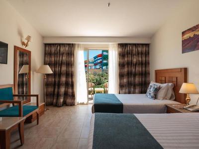 Hotelzimmer mit zwei Betten, Sesseln, Fernseher und Balkon mit Blick auf Außenanlage.