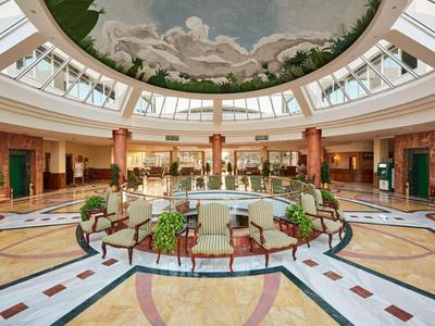 Světlé hotelové lobby s kruhovou odpočinkovou zónou, dekorativním malbou na stropě a přirozeným světlem přes velké světlíky.