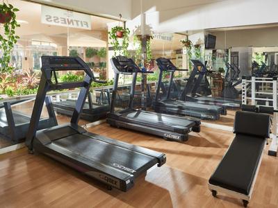 Moderní fitness s běžeckými pásy a činkami před velkými okny s rostlinami.